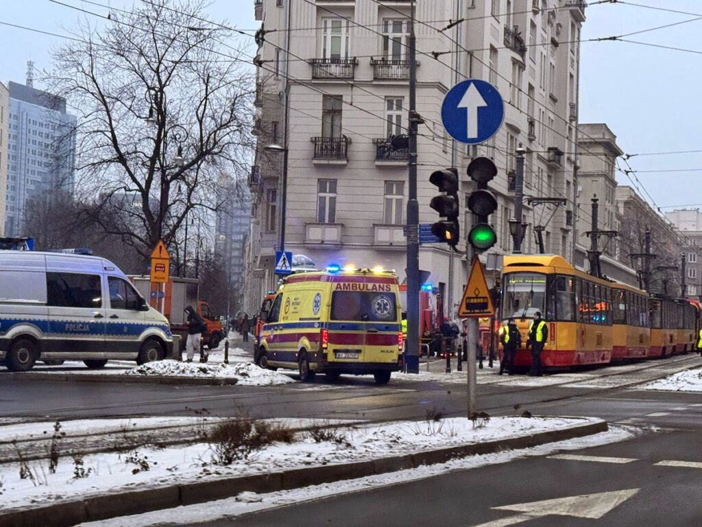 Karetka pogotowia oraz radiowóz policyjny na skrzyżowaniu, obok zatrzymany tramwaj w zimowej scenerii.