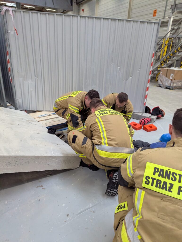 Czterech strażaków w uniformach pracuje nad uwolnieniem osoby uwięzionej pod betonową płytą w hali magazynowej.