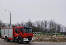 Czerwony wóz strażacki zatrzymany na skrzyżowaniu, w tle pociąg przejeżdżający obok zarośniętego terenu.