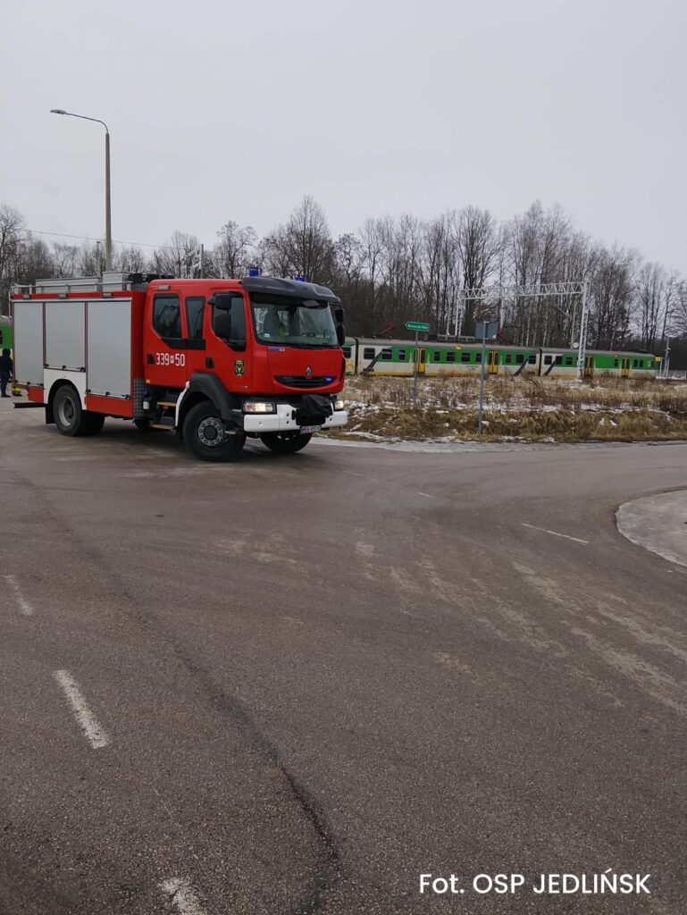 Czerwony wóz strażacki zatrzymany na skrzyżowaniu, w tle pociąg przejeżdżający obok zarośniętego terenu.