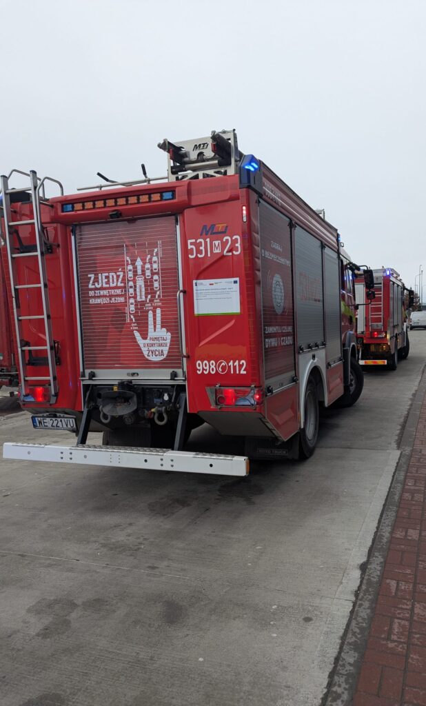 Czerwony wóz strażacki z włączonymi sygnałami świetlnymi zaparkowany na chodniku, obok drugiego wozu.