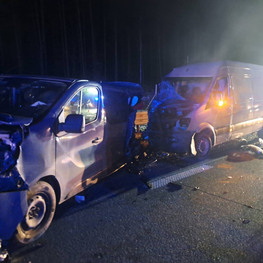 Dwa uszkodzone pojazdy dostawcze zderzone na drodze, otoczone przez policyjne światła w nocy.