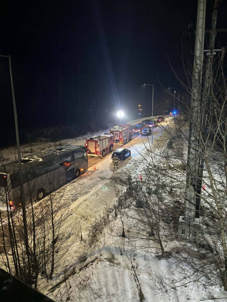 Na drodze w nocy stoją samochody, w tym autobus i pojazdy straży pożarnej, wokół których są włączone sygnały świetlne.