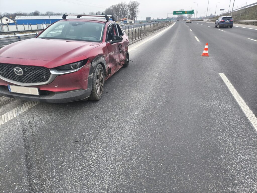 Czerwony samochód osobowy z uszkodzoną stroną stoi na pasie ruchu, obok znajduje się pomarańczowy trójkąt ostrzegawczy.
