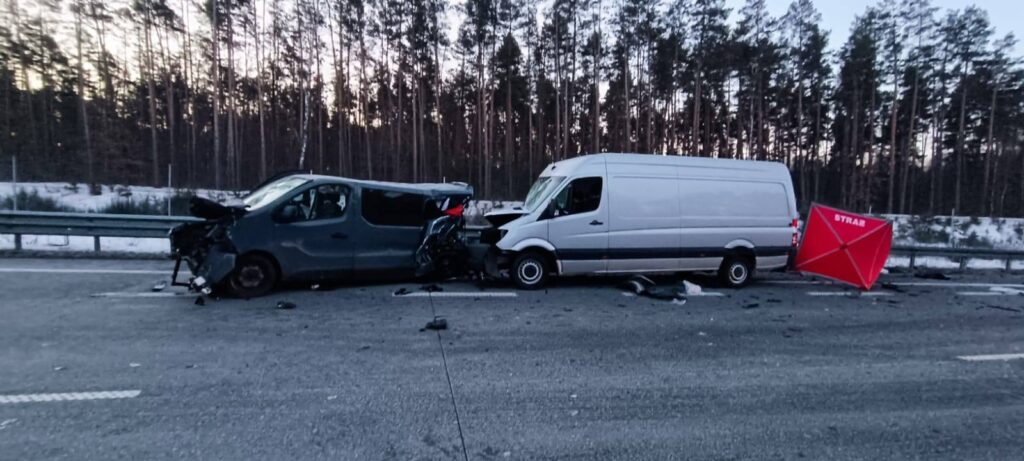 Dwa uszkodzone pojazdy dostawcze na drodze, obok leży czerwona tablica ostrzegawcza, w tle las.
