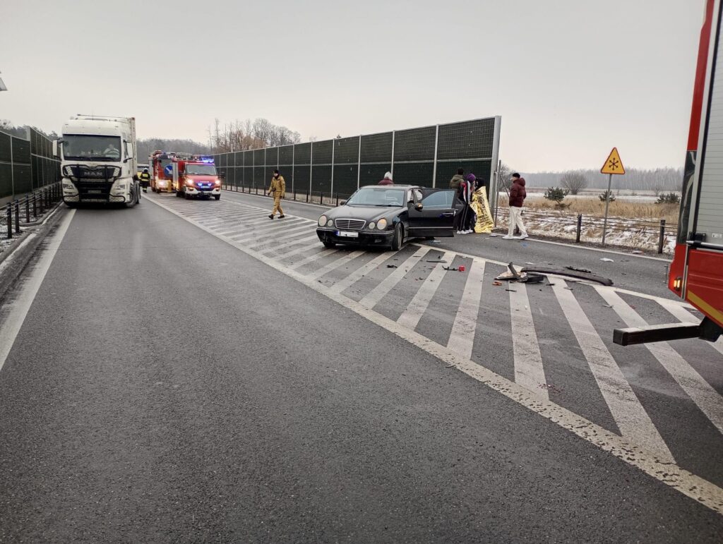 Na drodze stoi uszkodzony samochód osobowy, obok którego znajdują się strażacy oraz ratownicy medyczni. W tle widać ciężarówkę i samochód z włączonymi sygnałami alarmowymi. Na jezdni leżą fragmenty pojazdu.