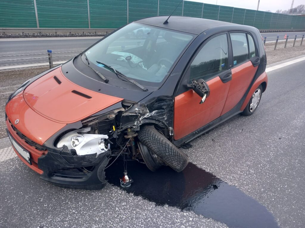Uszkodzony samochód osobowy z czarnym dachem i pomarańczowym nadwoziem stoi na poboczu drogi, z uszkodzoną przednią częścią i wyciekającymi płynami.