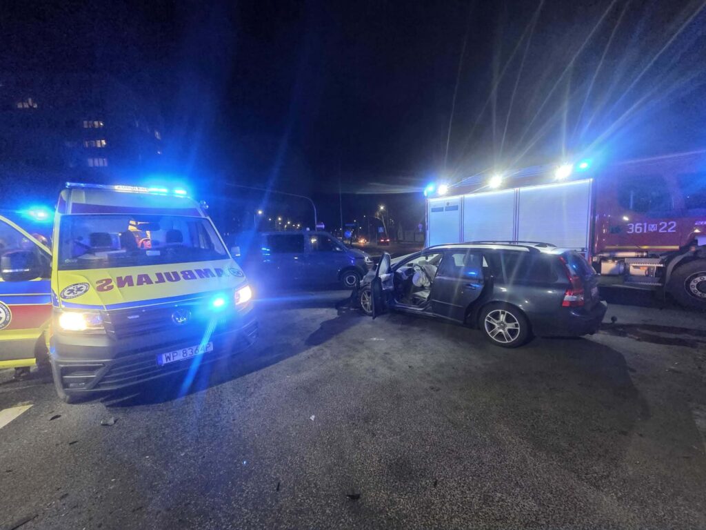 Ambulans z włączonymi sygnałami świetlnymi stoi obok uszkodzonego samochodu osobowego na drodze nocą.