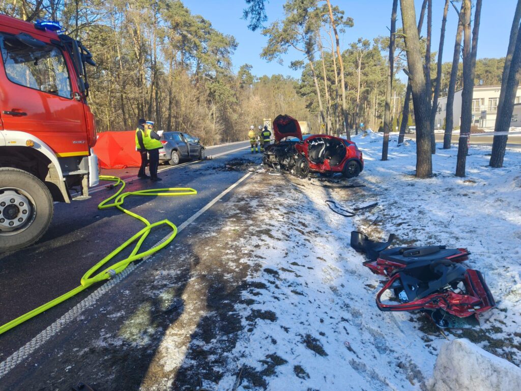Strażacy pracują nad wypadkiem drogowym, obok uszkodzonego pojazdu w lesie, w otoczeniu śniegu i drzew.