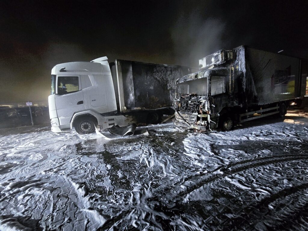 Dwa spalone ciężarówki na parkingu, pokryte pianą gaśniczą, w otoczeniu ciemnej nocy.