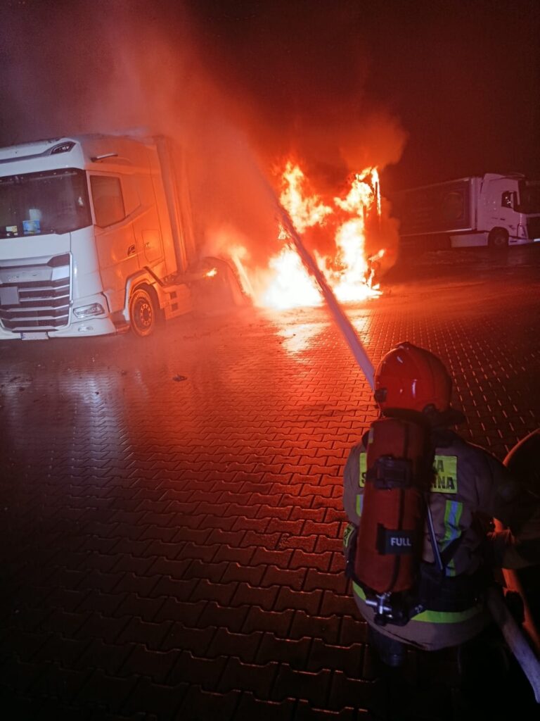 Strażak w hełmie gasi płonący samochód ciężarowy na nocnym parkingu, otoczony dymem i ogniem.