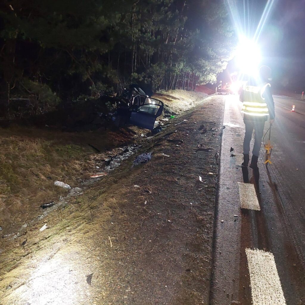 Policjant z latarką bada miejsce wypadku samochodowego obok drogi, gdzie leży zniszczony pojazd.