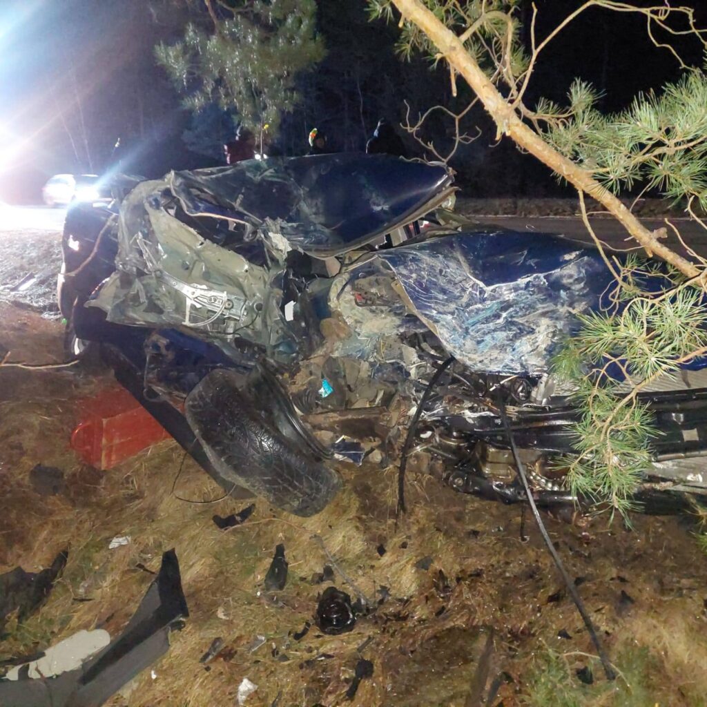 Zniszczony samochód leży na poboczu drogi, otoczony przez drzewa i resztki wraku.