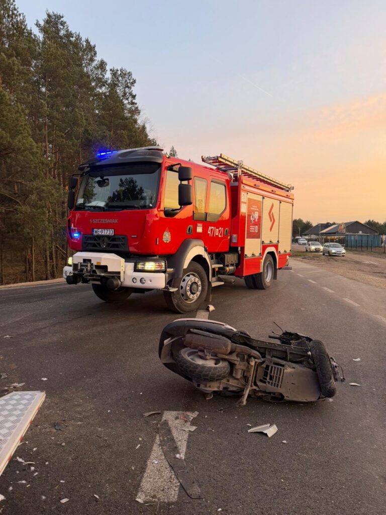 Czerwony wóz strażacki z włączonymi sygnałami świetlnymi stoi na drodze obok przewróconego skutera. W tle widać zarys drzew i budynków.