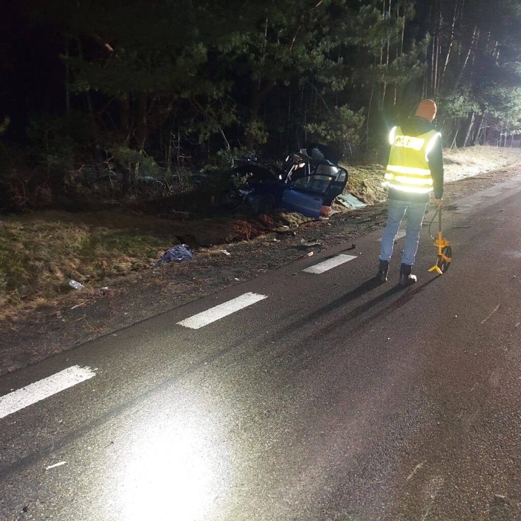 Mężczyzna w kamizelce odblaskowej bada miejsce wypadku obok drogi, gdzie stoi uszkodzony niebieski samochód.