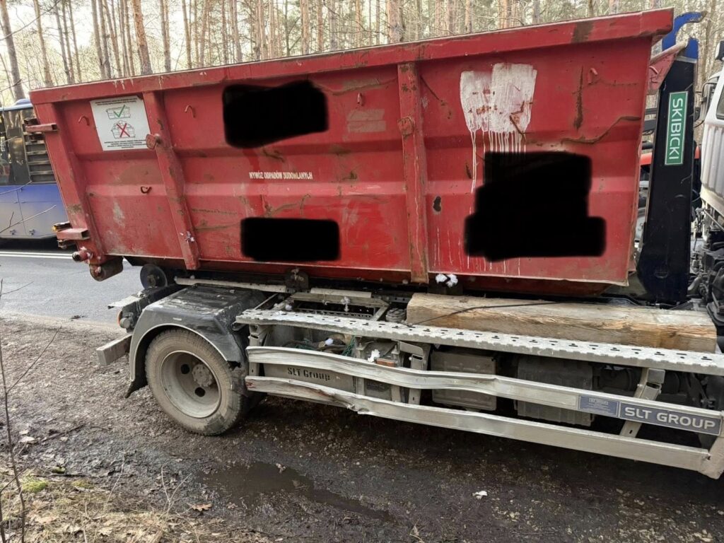 Czerwony kontener na śmieci umieszczony na przyczepie ciężarówki, stojący przy drodze w lesie.