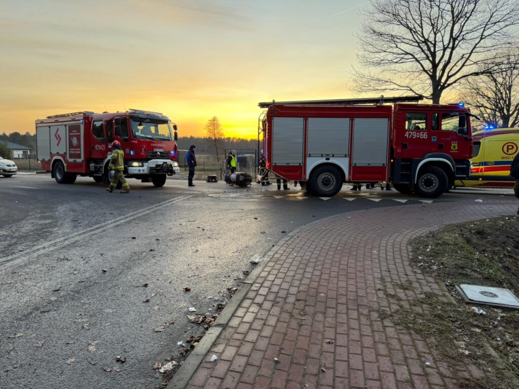 Strażacy pracują przy wypadku drogowym, obok dwóch wozów strażackich na skrzyżowaniu o zachodzie słońca.
