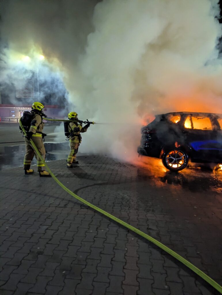 Dwaj strażacy w hełmach gaszą płonący samochód w gęstym dymie na placu.