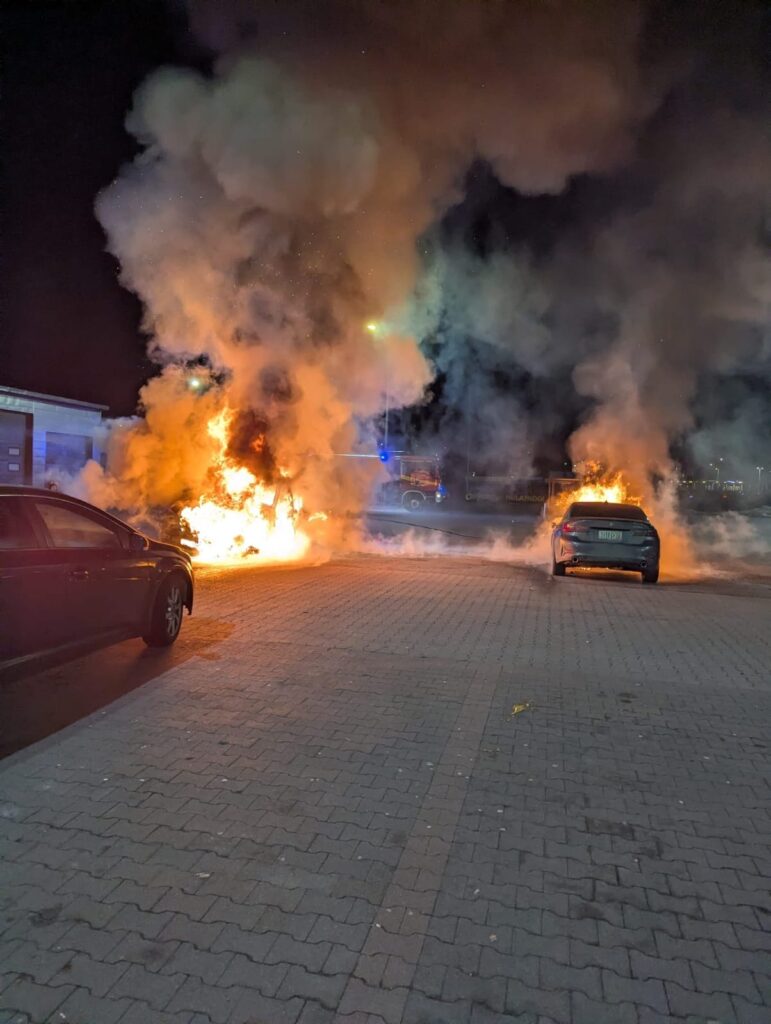 Pojazd płonący na parkingu, otoczony gęstym dymem i ogniem, w tle widoczne inne samochody.