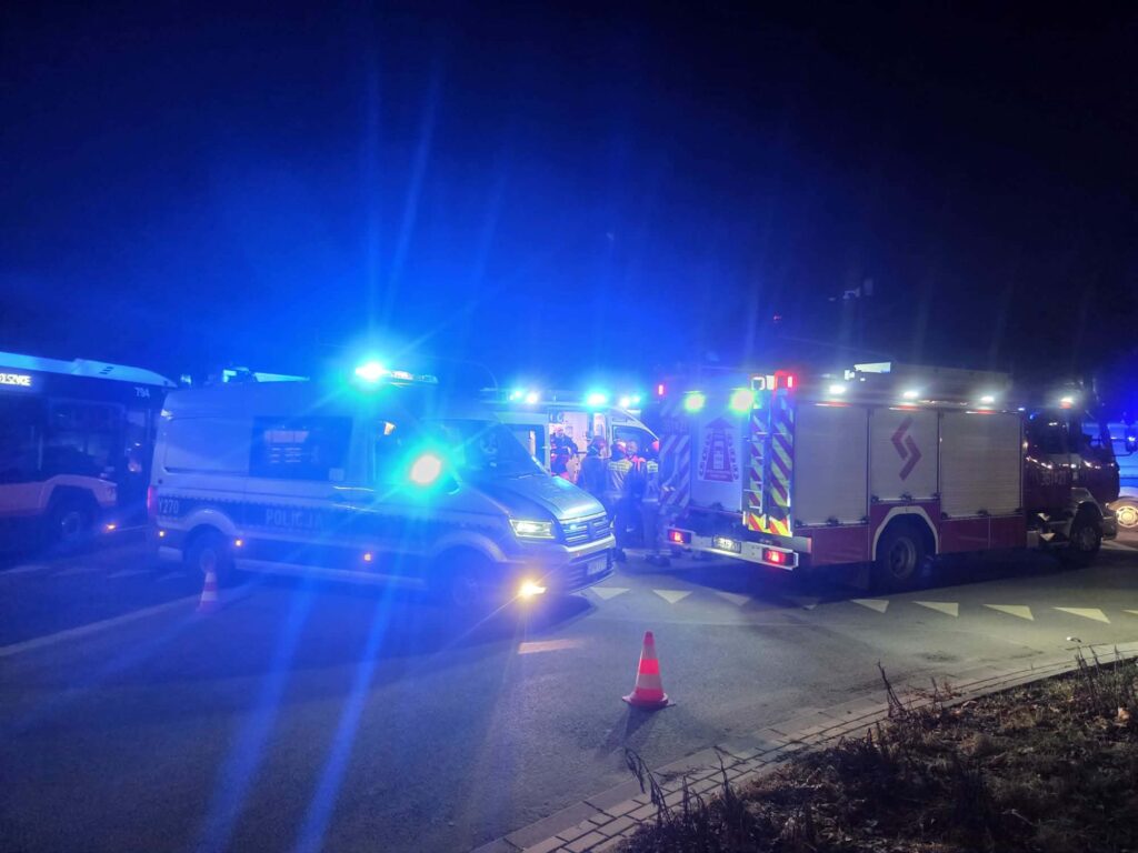 Karetka pogotowia i wóz strażacki na oświetlonej ulicy, w tle policja i zebrani ratownicy.