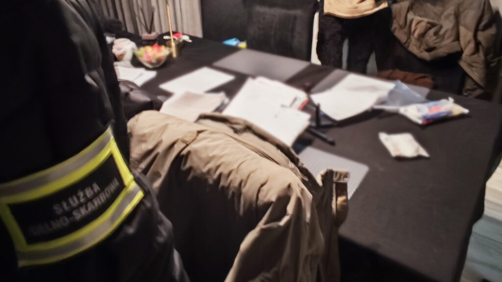 Funkcjonariusz w mundurze z napisem "Służba Celno-Skarbowa" stoi przy stole z dokumentami i notatkami.