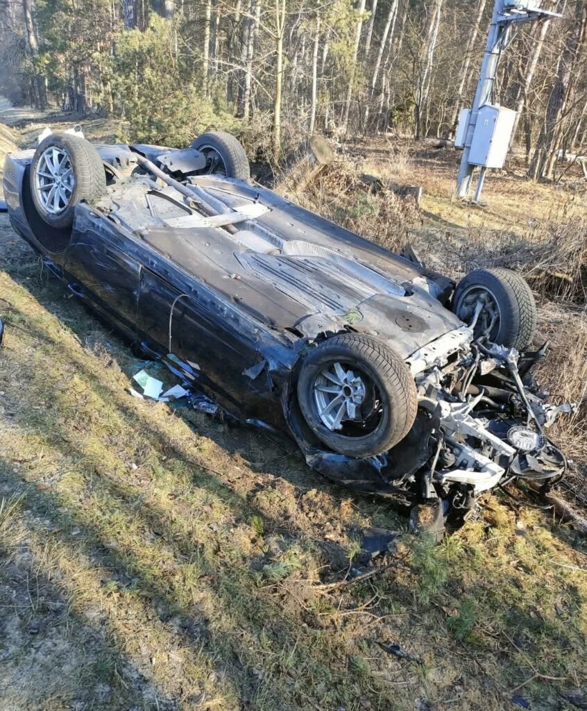 Czarny samochód osobowy leży na dachu na poboczu leśnej drogi, otoczony przez drzewa i trawę.