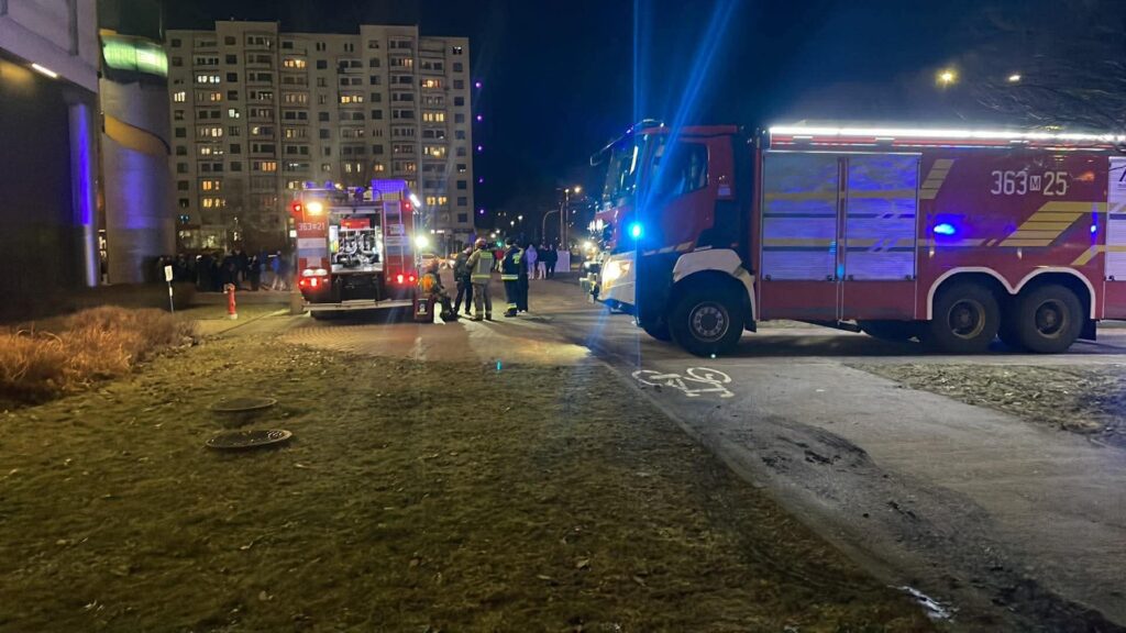 Strażacy rozmawiają obok wozu strażackiego na oświetlonej ulicy, w tle grupa ludzi oraz budynki mieszkalne.
