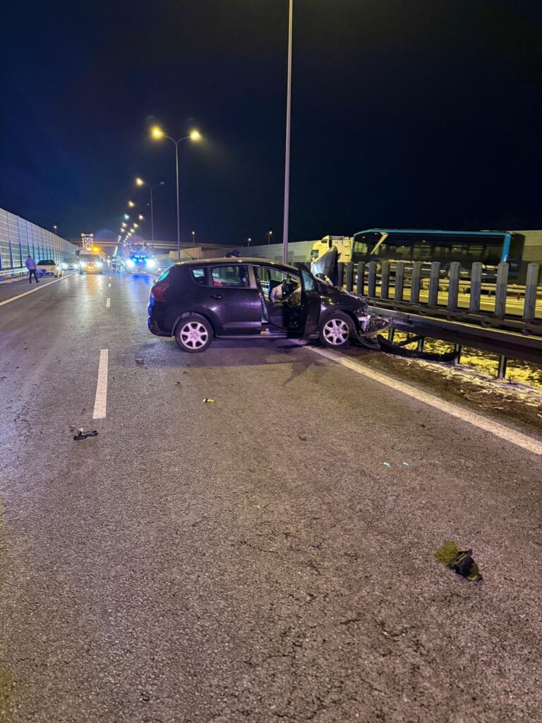 Czarny samochód osobowy z otwartymi drzwiami stoi na pasie ruchu, obok widać pojazdy służb ratunkowych.