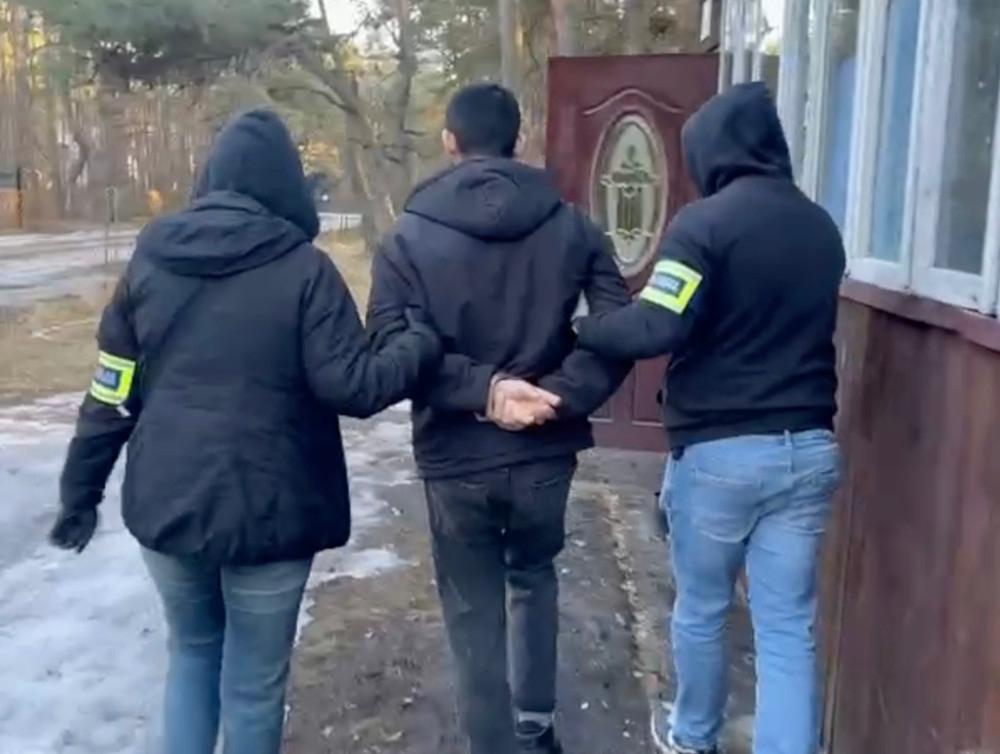 Dwóch funkcjonariuszy w czarnych kurtkach eskortuje mężczyznę z rękami z tyłu w lesie.