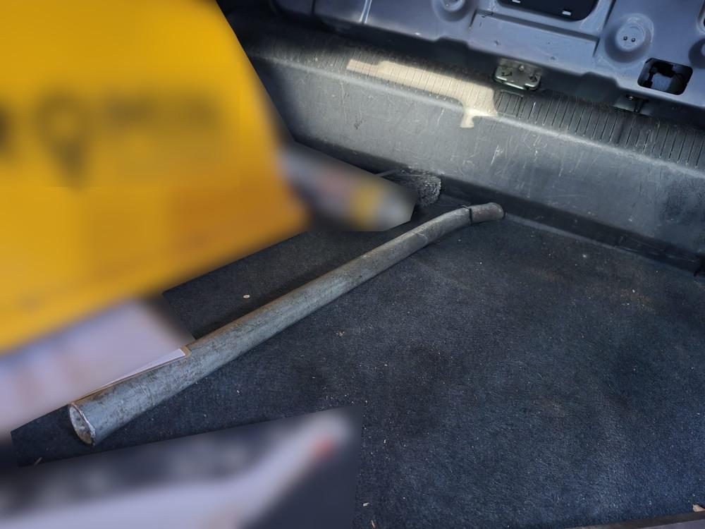 Metalowa rura leżąca na wykładzinie bagażnika pojazdu, obok fragmentów dokumentów.