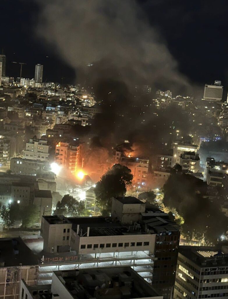 W tle miasta widać pożar, z którego unoszą się kłęby czarnego dymu, a w oddali widać płomienie.