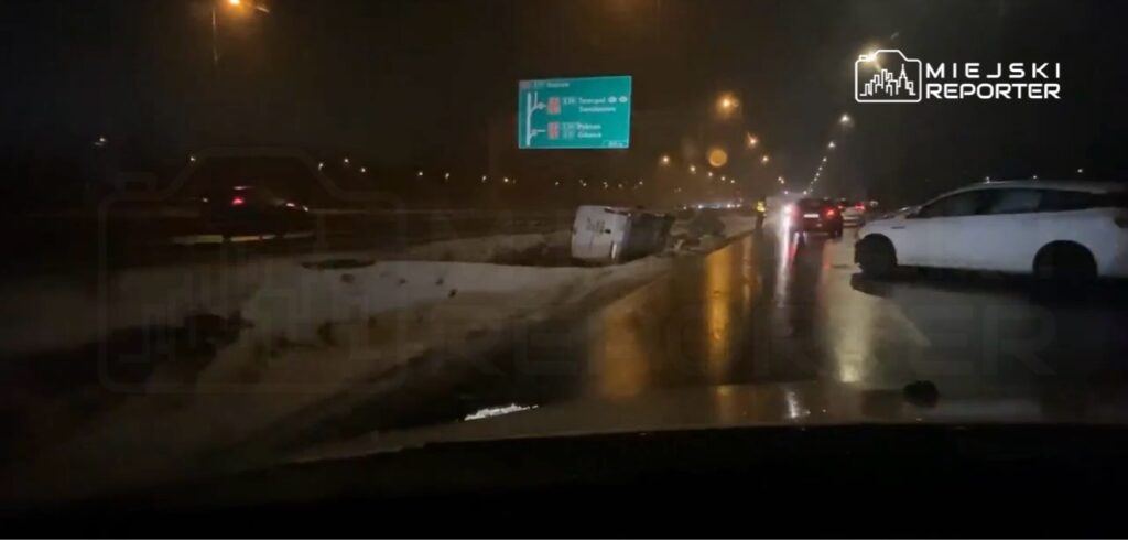 Samochód leżący na boku na mokrej jezdni, obok znaki drogowe i przejeżdżające pojazdy w nocy.