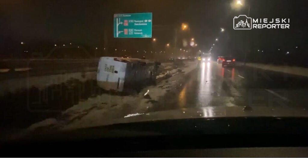 Przewrócony pojazd leży na poboczu drogi, obok znaki kierunkowe, w nocy przy deszczowej pogodzie.