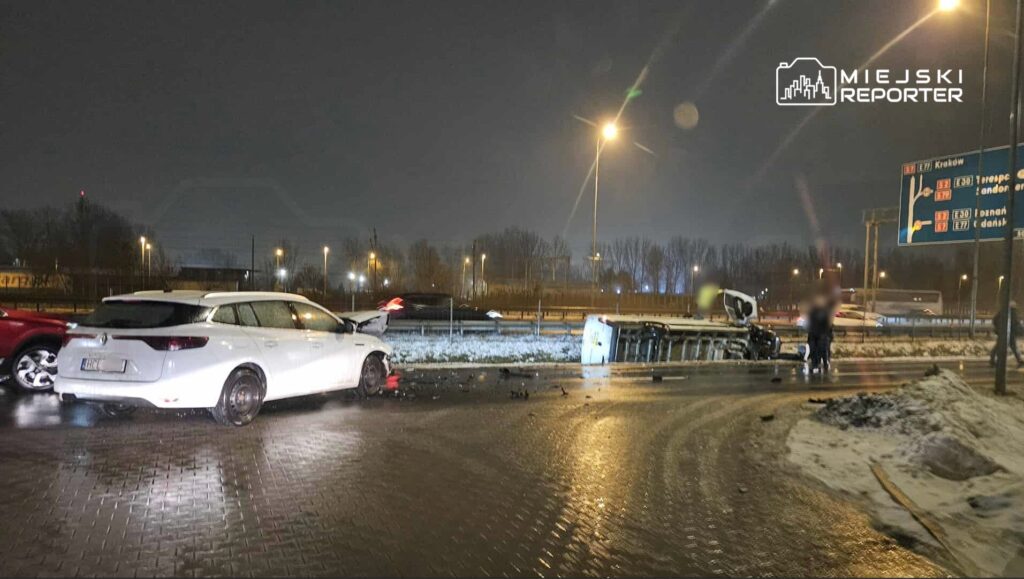 Zderzenie dwóch samochodów na ośnieżonej drodze, jeden z pojazdów przewrócony na bok, w tle znaki drogowe.