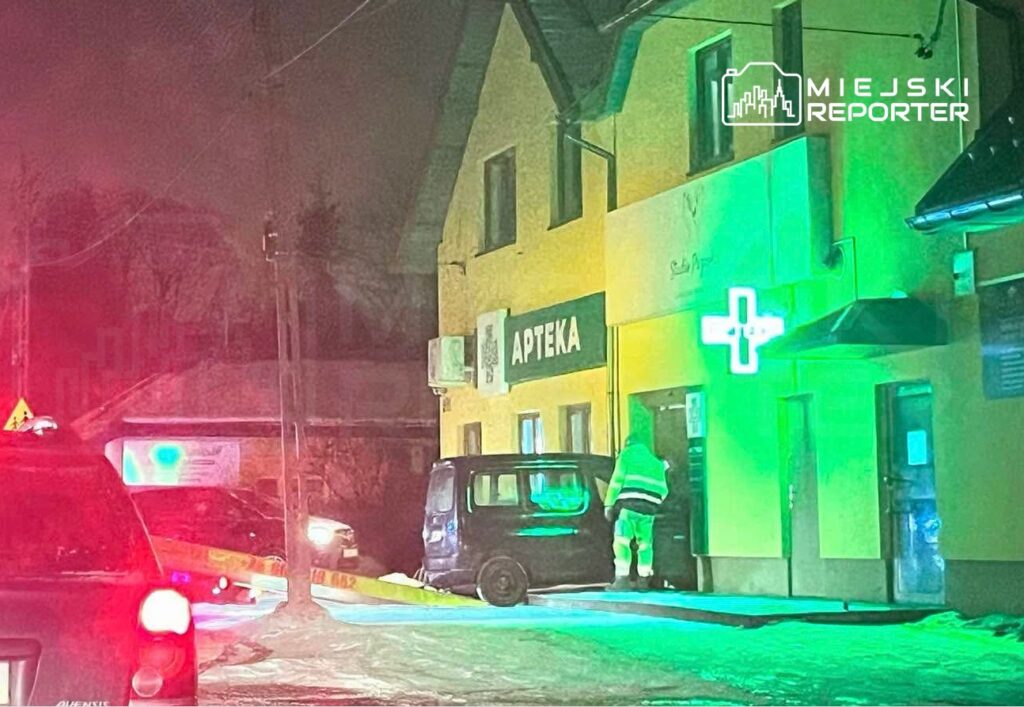 Mężczyzna w odblaskowej kamizelce podchodzi do apteki, obok zaparkowany niebieski samochód, nocna sceneria.