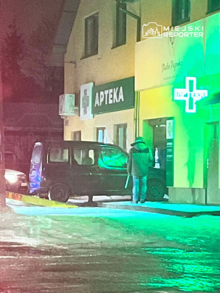 Mężczyzna w szarej kurtce stoi obok ciemnozielonego samochodu przed apteką, oświetloną neonowym znakiem.