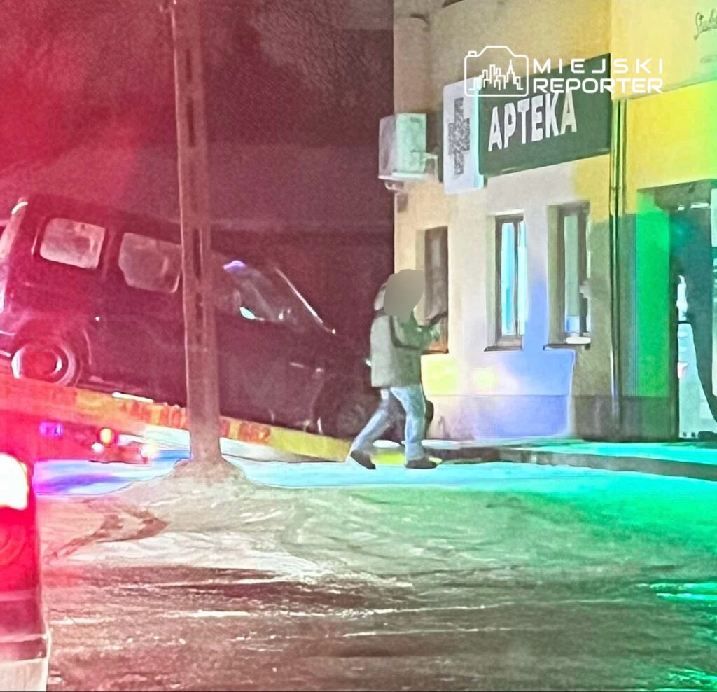 Czarny samochód osobowy wciągany na lawetę obok apteki w nocy, w tle widoczne migające światła policyjne.