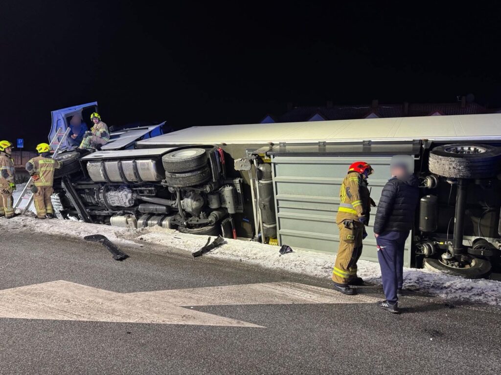 Strażacy pracują przy przewróconym samochodzie ciężarowym na drodze w nocy, obok leży uszkodzony fragment naczepy.