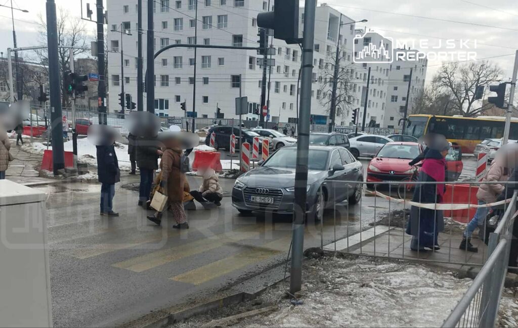 Grupa osób na pasach, jedna osoba siedzi na ziemi, samochody zatrzymane, w tle nowoczesne budynki mieszkalne.
