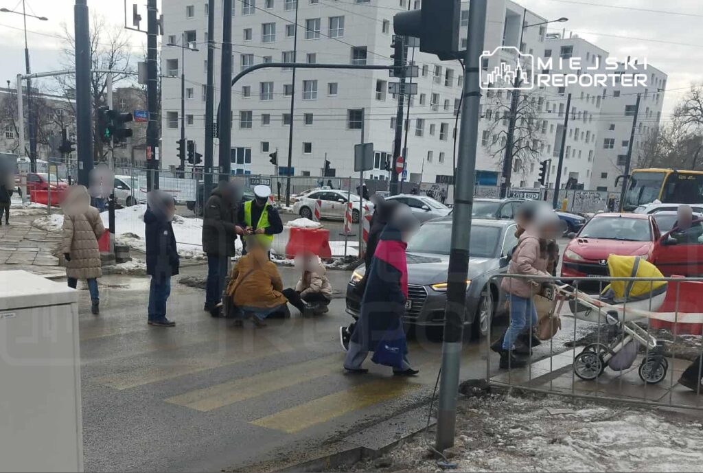 Grupa osób zgromadzona przy przejściu dla pieszych, gdzie mężczyzna w kamizelce odblaskowej pomaga osobie siedzącej na ziemi. W tle widoczne samochody i nowoczesne budynki.