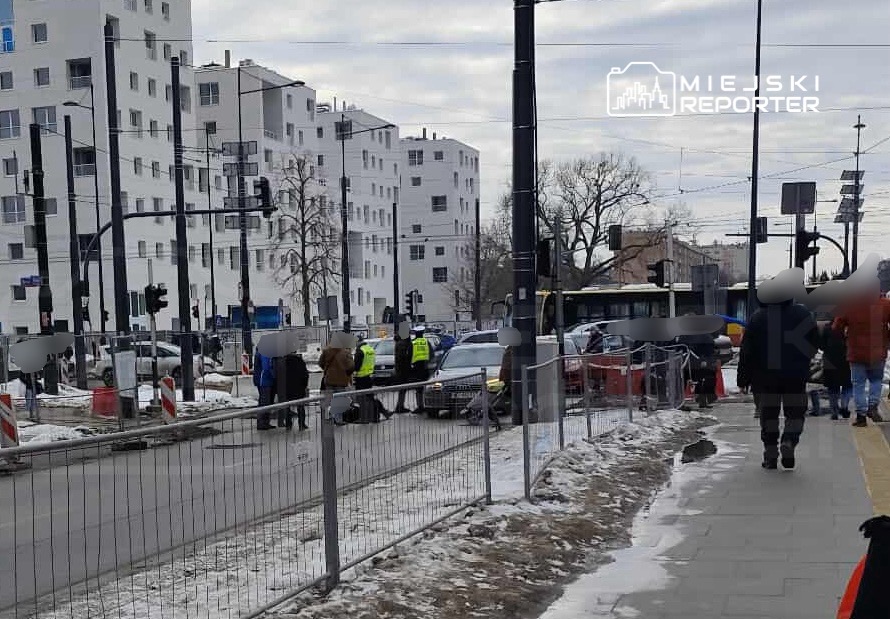 Grupa policjantów bada miejsce wypadku drogowego, w tle nowoczesne budynki i ruch uliczny.