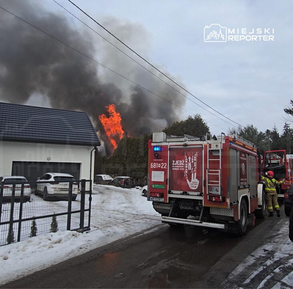 Strażacy przy wozie strażackim gaszą pożar w pobliżu domów, z dużymi płomieniami i dymem unoszącym się w tle.