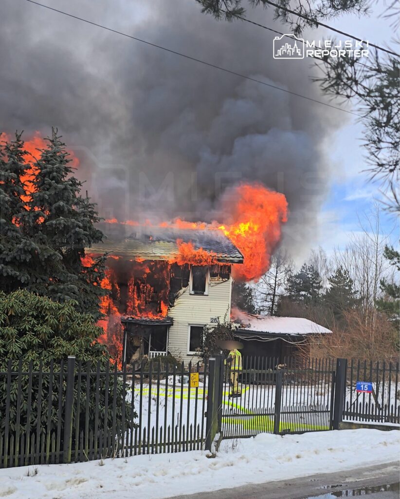 Dom płonie, wydobywają się płomienie i gęsty dym, a strażak podchodzi do budynku w zimowym otoczeniu.