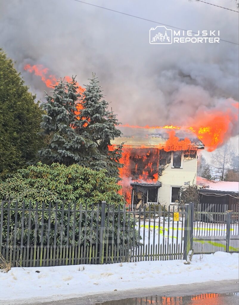 Dom w płomieniach z intensywnym ogniem i dymem, otoczony zielonymi drzewami i ogrodzeniem.