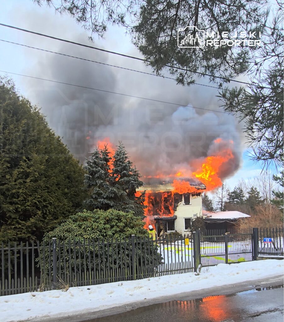Dom płonie, wydobywa się z niego gęsty dym oraz płomienie, w otoczeniu śniegu i zieleni drzew.