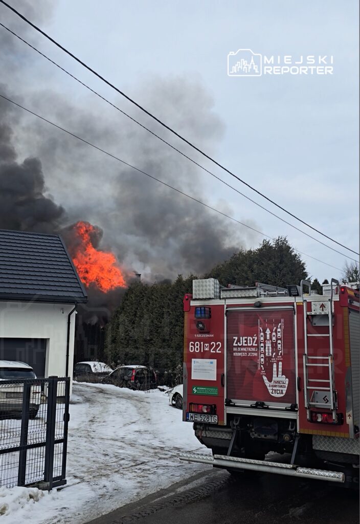 Pożar w budynku, z którego wydobywa się dym i płomienie, w tle widać wóz strażacki na śnieżnej drodze.