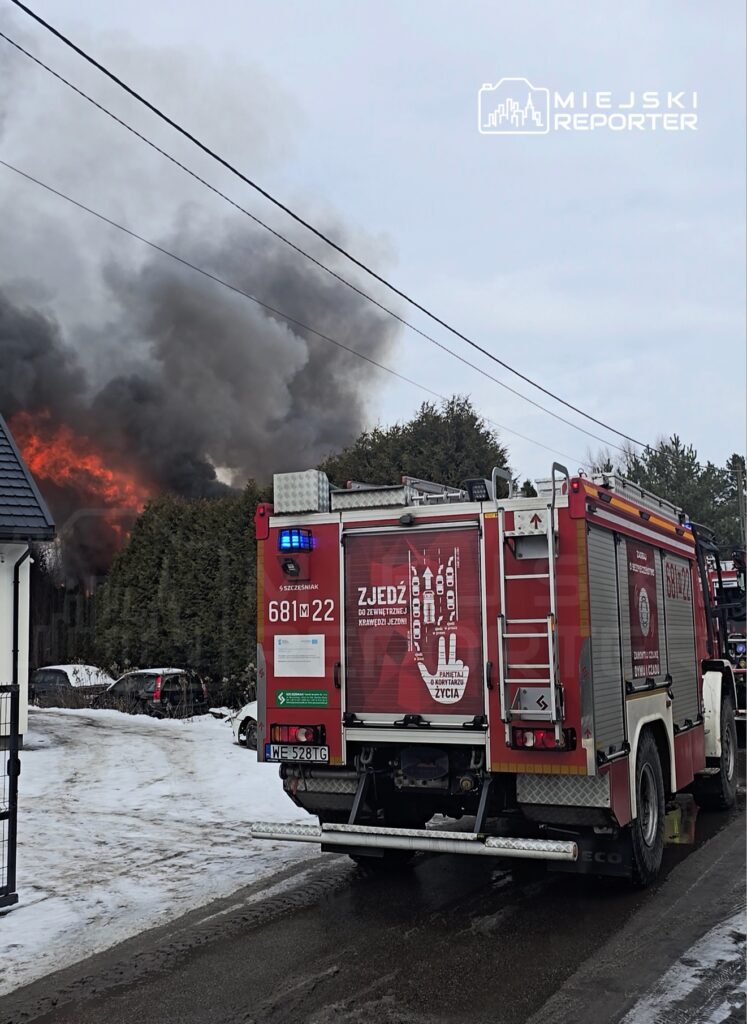 W tle widoczne płomienie i gęsty dym unoszący się z budynku, z przodu widać wóz strażacki z włączonymi sygnałami świetlnymi.