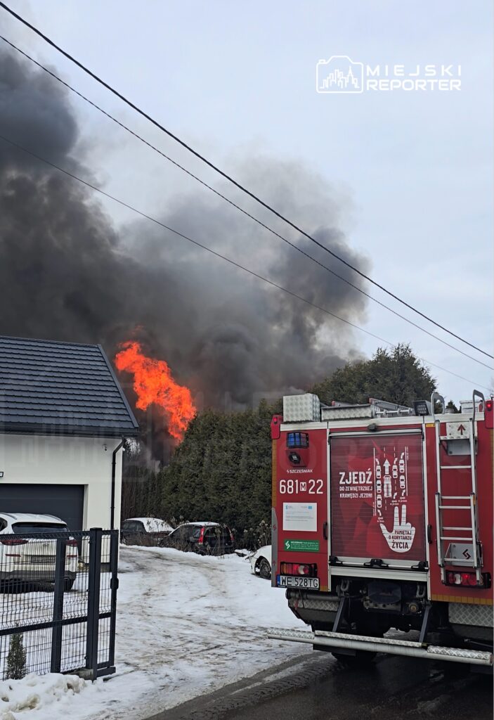 Pożar w okolicy, z płomieniami i dużą ilością dymu. W tle widać wóz strażacki i zaśnieżoną ulicę.