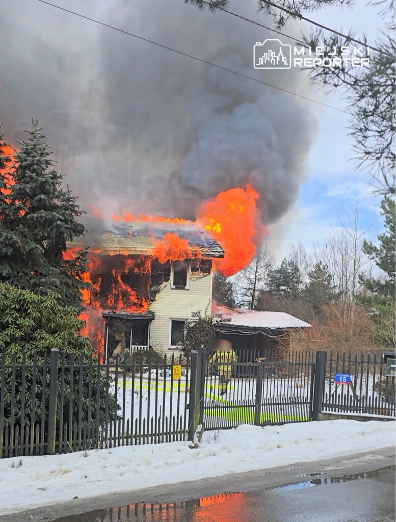 Dom w płomieniach, z którego wydobywa się gęsty dym, otoczony śniegiem i ogrodzeniem.