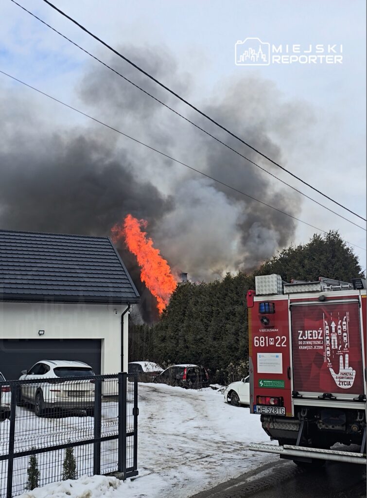 Pożar wydobywający się z dachu budynku, w tle gęsty dym i ogień, w otoczeniu śniegu i zaparkowanych samochodów.
